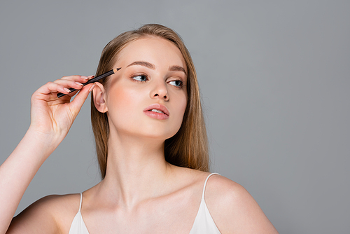 young woman applying brown eyebrow 펜슬 and looking away isolated on grey