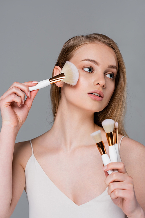 young woman holding set of different cosmetic brushes and applying face powder isolated on grey