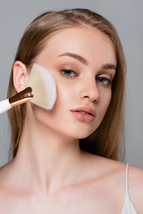 young woman holding cosmetic brush and applying face powder isolated on grey