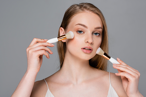 young woman holding different cosmetic brushes and applying face powder isolated on grey