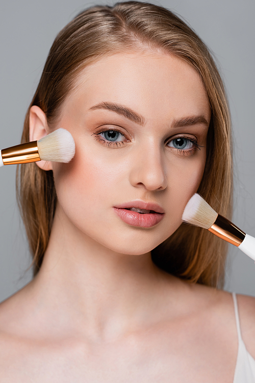 close up of young woman holding different cosmetic brushes and applying face powder isolated on grey
