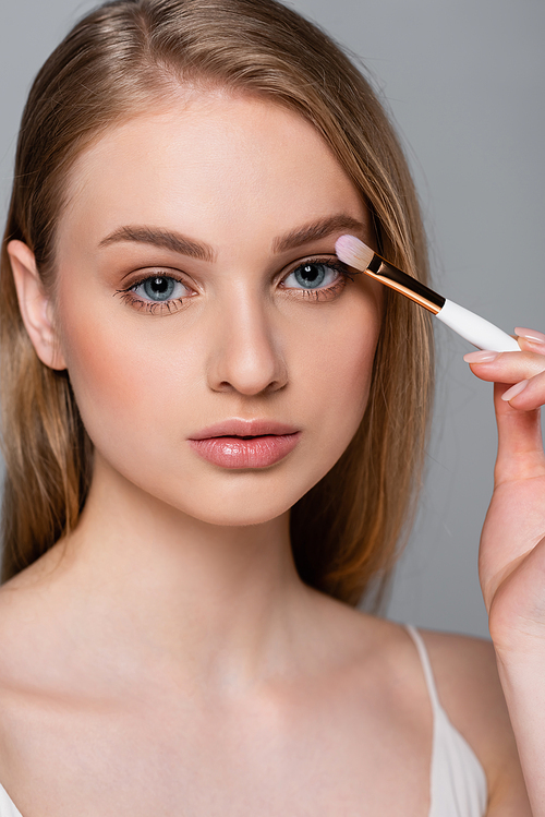pretty young woman holding cosmetic brush and applying eye shadow isolated on grey