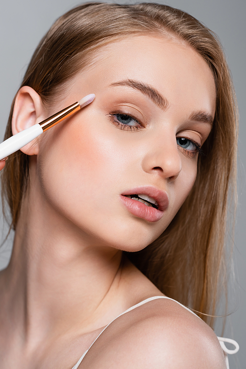 close up of young woman holding cosmetic brush and applying eye shadow isolated on grey