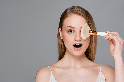 amazed young woman holding cosmetic brush isolated on grey