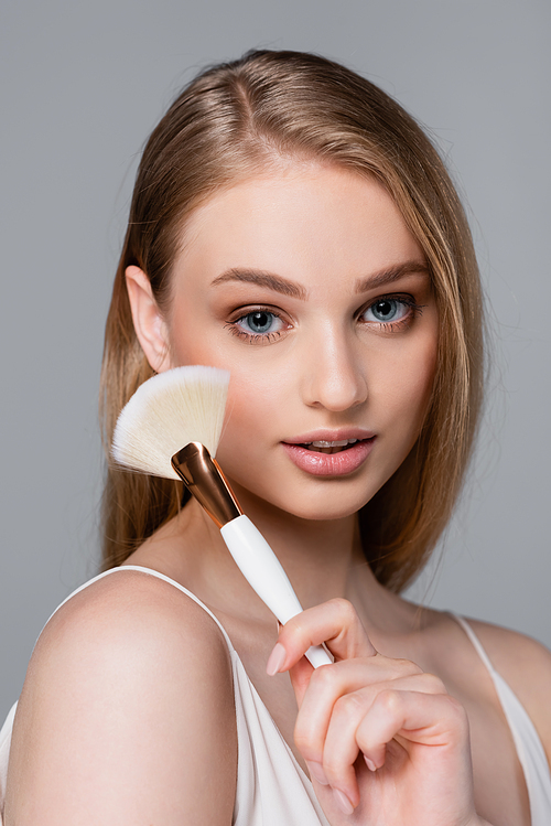 young and pretty woman holding cosmetic brush isolated on grey