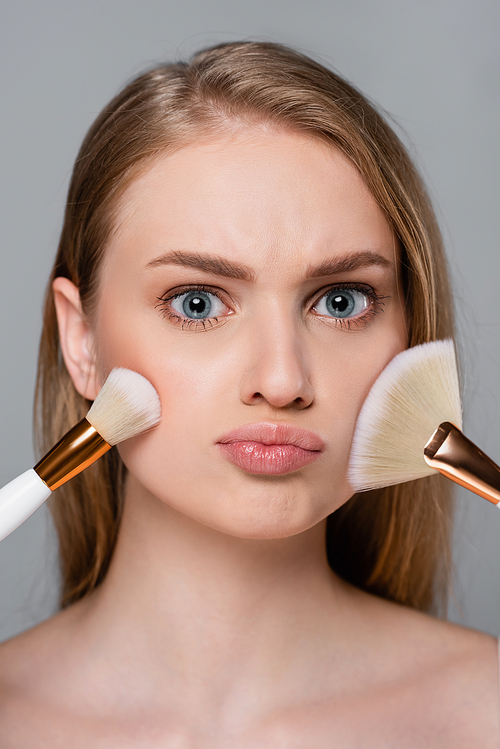 young woman holding cosmetic brushes and applying face powder isolated on grey