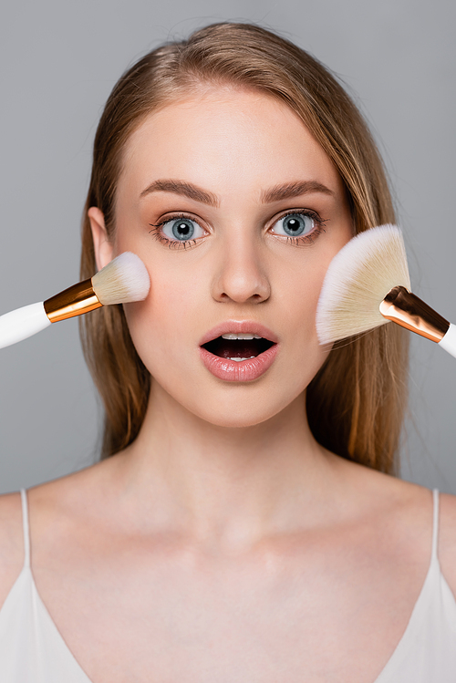 amazed young woman holding cosmetic brushes near face isolated on grey