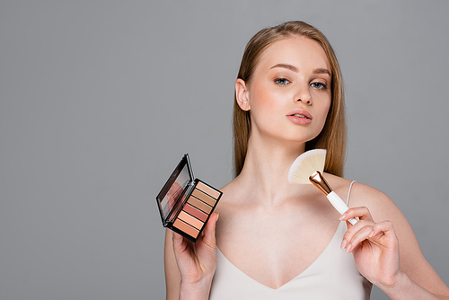 pretty woman holding cosmetic brush and eye shadow and blush palette isolated on grey