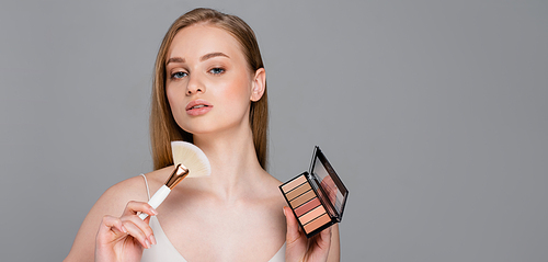 young woman holding cosmetic brush and eye shadow and blush palette isolated on grey, banner