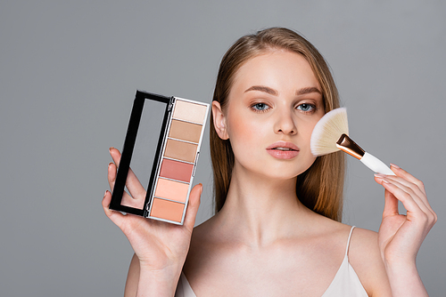 young woman holding cosmetic brush and eye shadow and blush palette isolated on grey