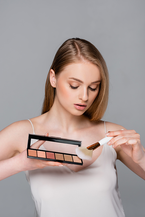 young woman holding cosmetic brush applying blush from palette isolated on grey