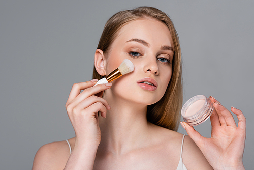 young woman holding cosmetic brush while applying face powder and container isolated on grey