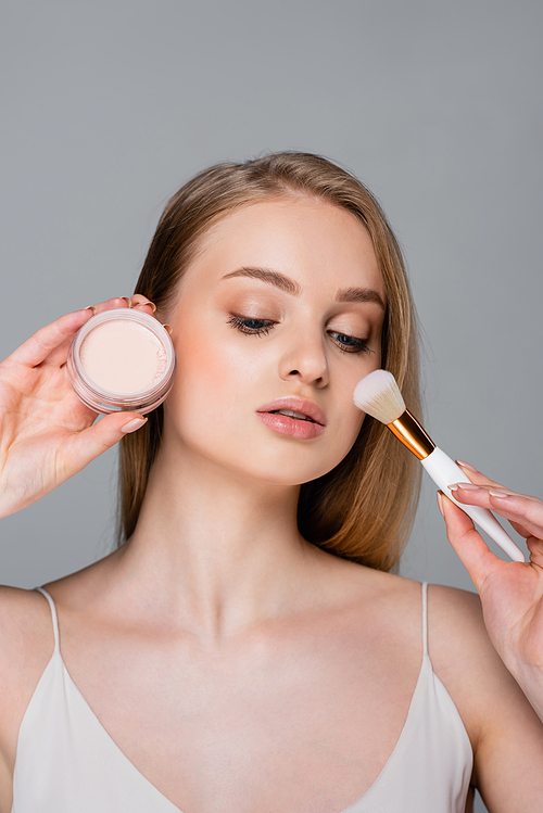 young woman holding cosmetic brush and face powder container isolated on grey