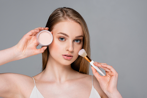 young woman holding cosmetic brush and face powder isolated on gray