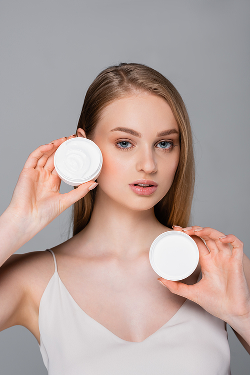 pretty young woman holding container with cosmetic cream isolated on grey