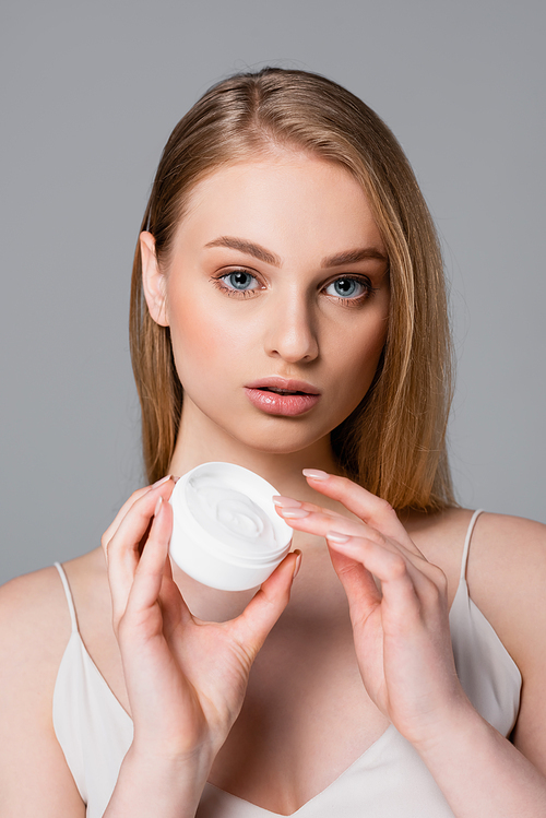 pretty young woman holding container with cosmetic cream isolated on gray