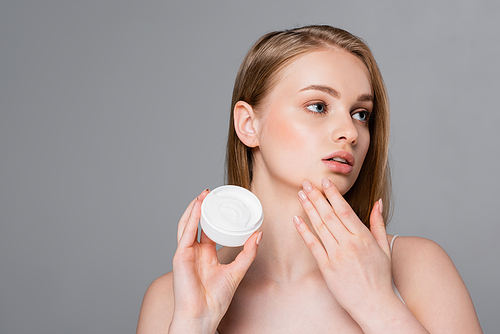 pretty young woman holding container with cosmetic cream and looking away isolated on grey