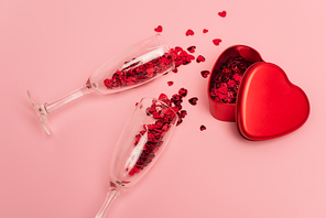 high angle view of champagne glasses with confetti hearts near metallic heart-shaped box on pink
