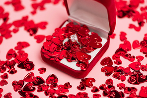 close up of jewelry box with diamond ring near red confetti hearts on pink