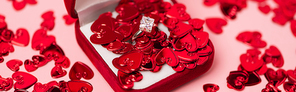 close up of jewelry box with diamond ring near red confetti hearts on pink, banner