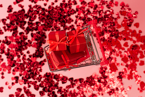top view of small shopping cart with presents near shiny red confetti hearts on pink