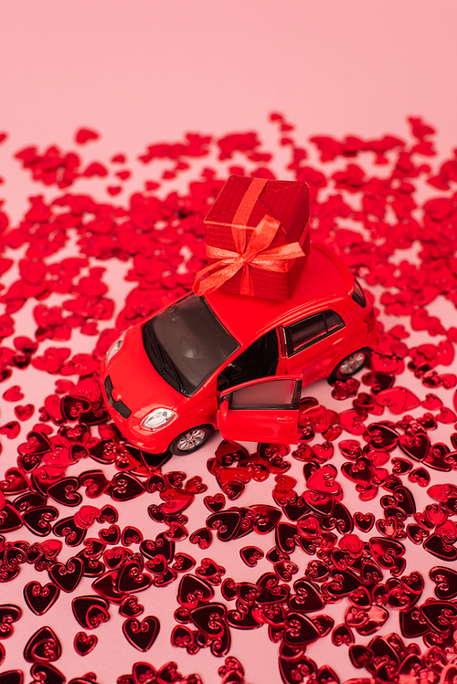 high angle view of toy car with wrapped present near shiny red confetti hearts on pink