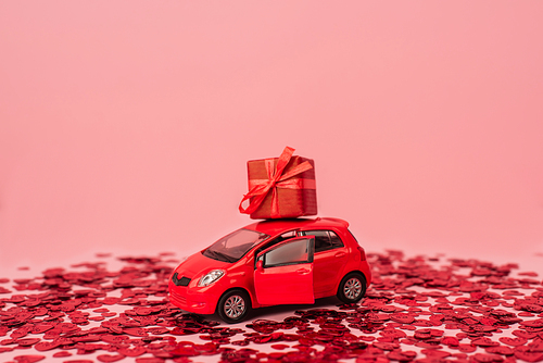 toy car with wrapped present near shiny red confetti hearts isolated on pink