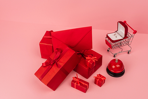high angle view of red wrapped gift boxes and envelope near shopping cart with engagement ring on pink