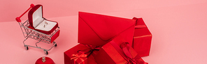 red wrapped gift boxes and envelope near shopping cart with engagement ring on pink, banner