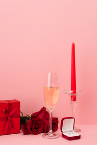 glass of champagne near gift box, roses, engagement ring and candle on pink