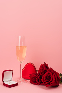 glass of champagne near heart-shaped box, roses and engagement ring on pink