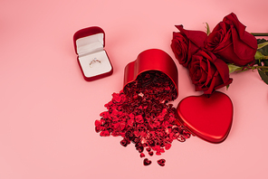 high angle view of metallic heart-shaped box with confetti near red roses and engagement ring on pink