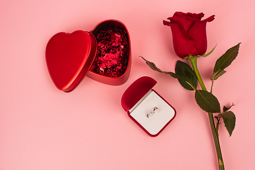 top view of red rose near box with heart-shaped confetti and engagement ring on pink