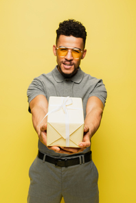 blurred african american man in sunglasses and grey tennis shirt holding wrapped present isolated on yellow