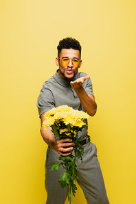 stylish african american man in sunglasses holding bouquet of flowers and sending air kiss isolated on yellow