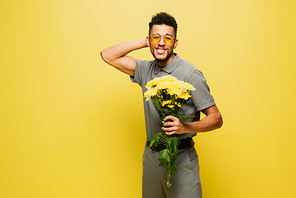 happy african american man in sunglasses holding bouquet of flowers on yellow