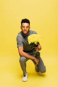 african american man in sunglasses holding bouquet of flowers and standing on knee on yellow