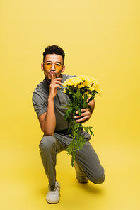 african american man in sunglasses holding bouquet of flowers while standing on knee and showing hush sign on yellow