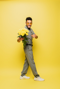 full length of happy african american man in sunglasses holding bouquet of flowers and standing on yellow