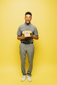 full length of excited african american man in sunglasses and grey tennis shirt holding retro telephone on yellow