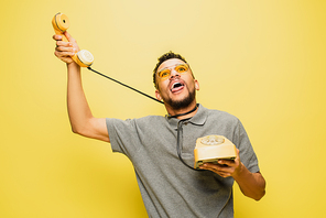 young african american man in sunglasses sticking out tongue while choking himself with telephone cable on yellow