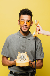 woman holding handset near african american man in sunglasses and grey tennis shirt  isolated on yellow