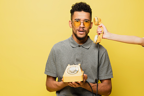 woman holding handset near shocked african american man in sunglasses and grey tennis shirt on yellow