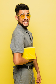 happy african american man in sunglasses and grey tennis shirt holding book isolated on yellow