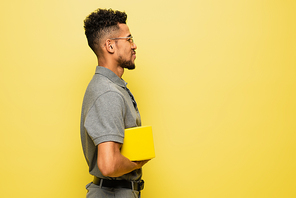 side view of happy african american man in sunglasses and grey tennis shirt holding book isolated on yellow