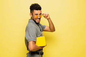 young african american man adjusting sunglasses and holding book on yellow