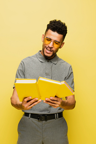 happy african american man in sunglasses and grey tennis shirt holding books isolated on yellow