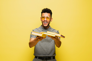 cheerful african american man in sunglasses and grey tennis shirt holding books on yellow