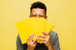 african american man in sunglasses covering face with books isolated on yellow