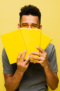 african american man in sunglasses covering face while holding books isolated on yellow